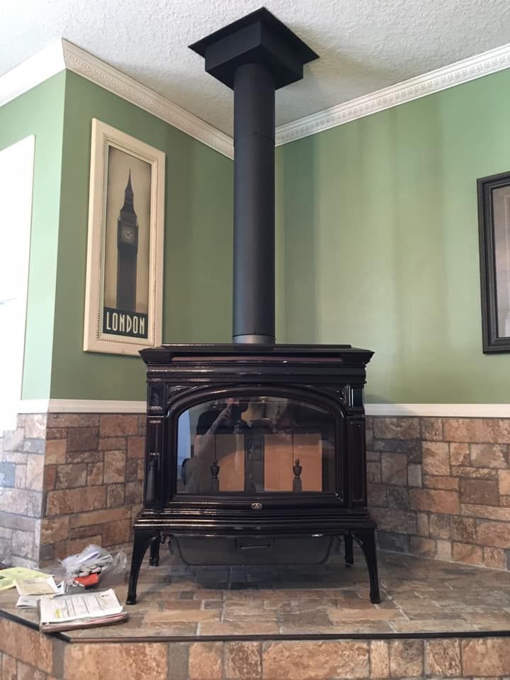 A black wood-burning stove with a black chimney pipe against green walls, a stone-like base, and artwork.