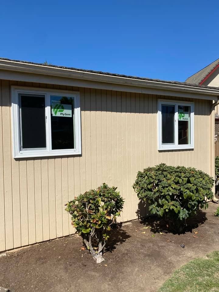 Exterior view of a beige building with two white-framed windows, two green bushes, and blue sky.
