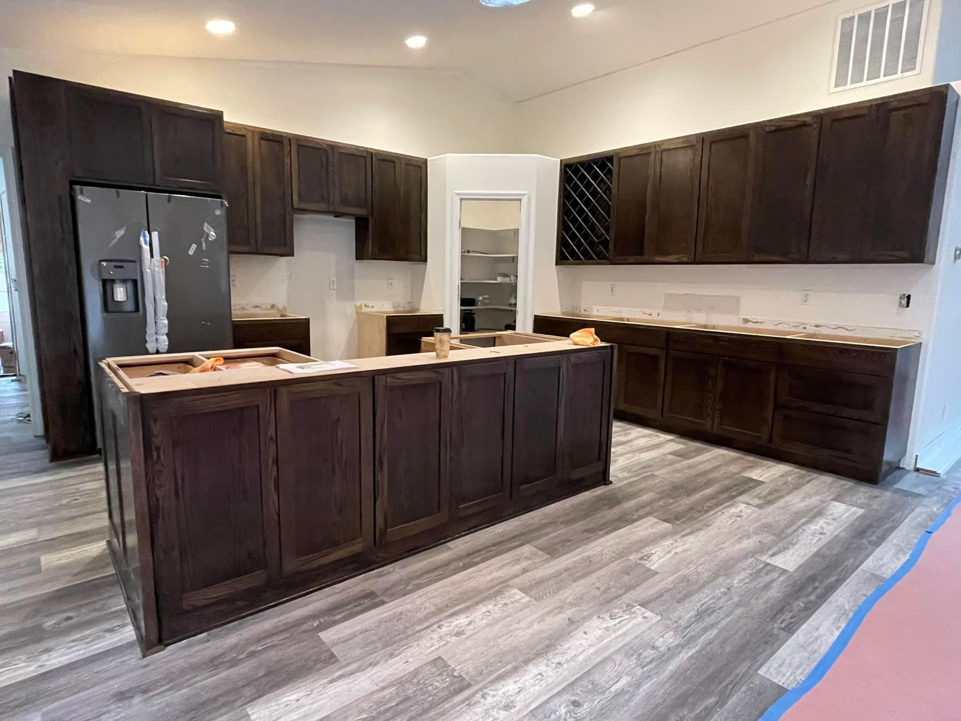 Kitchen cabinets being installed; dark wood, island, grey flooring, refrigerator.