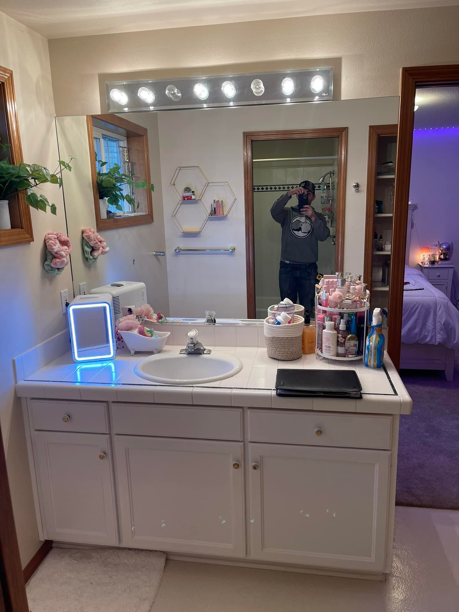 Bathroom with white vanity, mirror reflecting person taking a photo. Contains decor, lights and open door to bedroom.