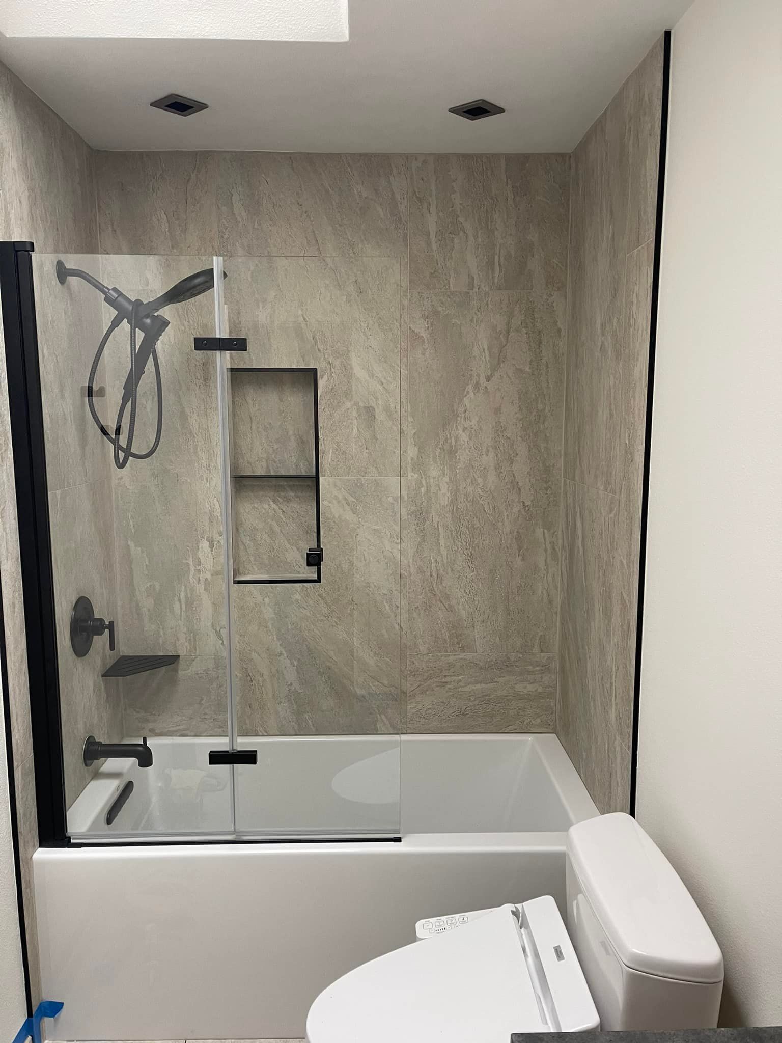 A modern bathroom with a bathtub, shower, and toilet. Gray tile walls and black fixtures.