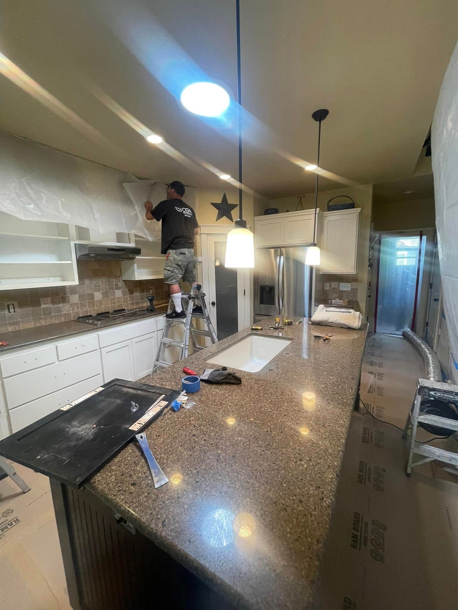 Person on ladder working on kitchen backsplash, surrounded by covered countertops and cabinets.