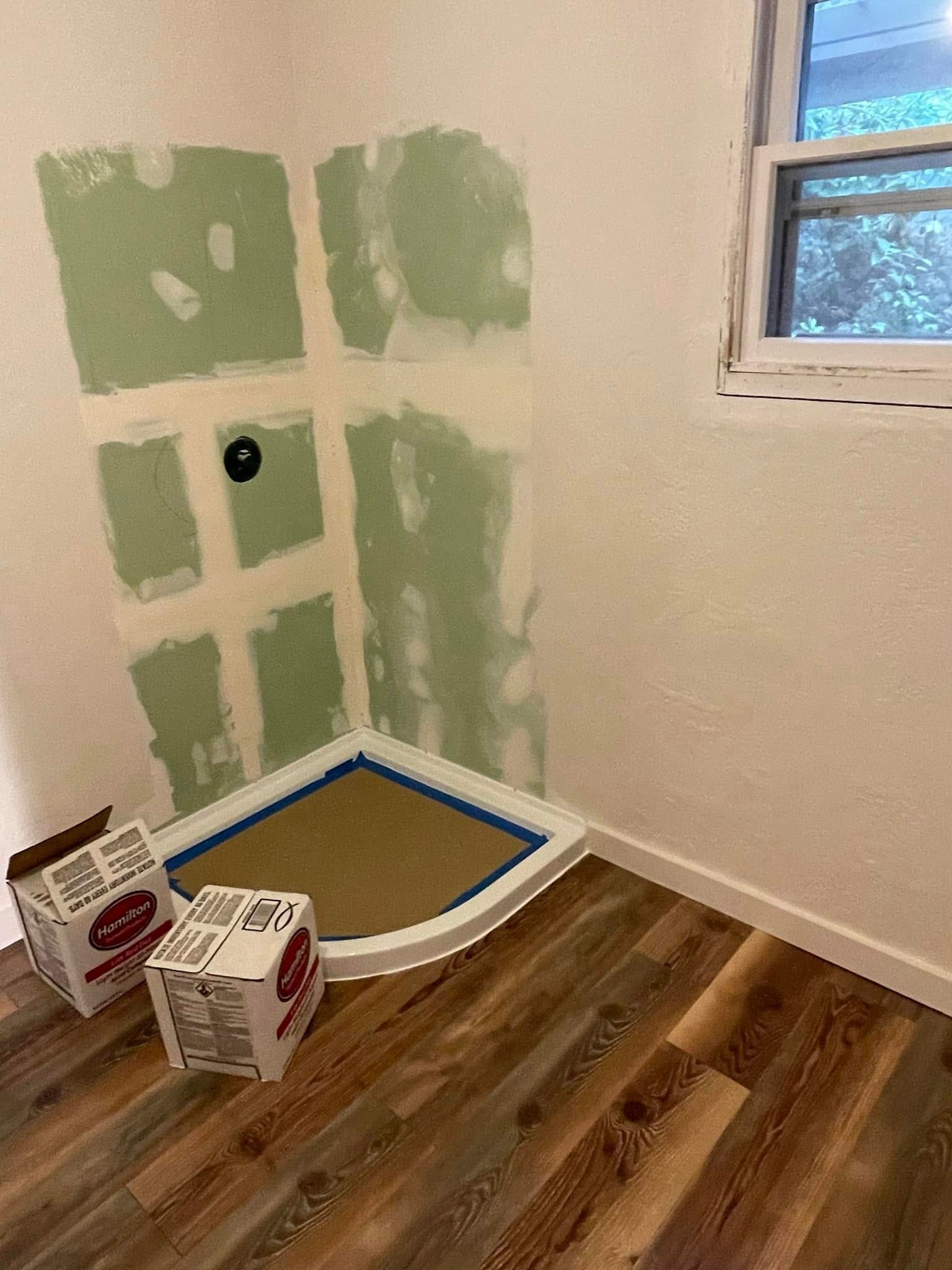 Bathroom renovation. Green wall, new shower base, and boxes sit on wood-look flooring near a window.