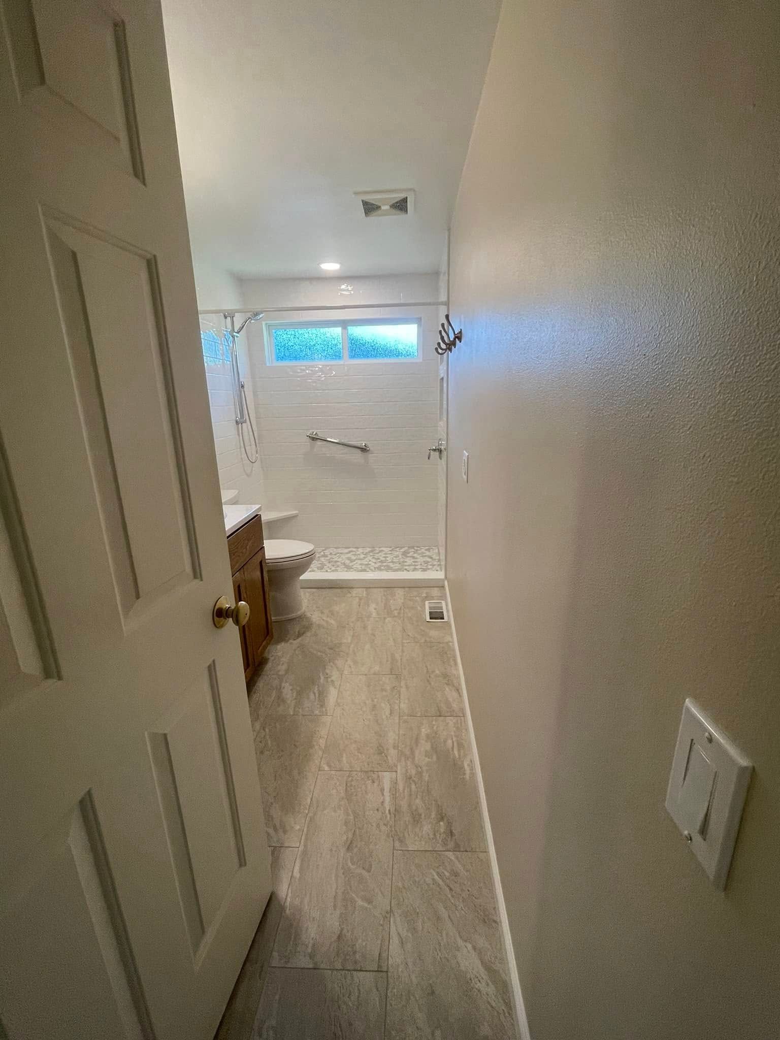 Bathroom with gray tile floor, white shower, wooden vanity, and door on the left.