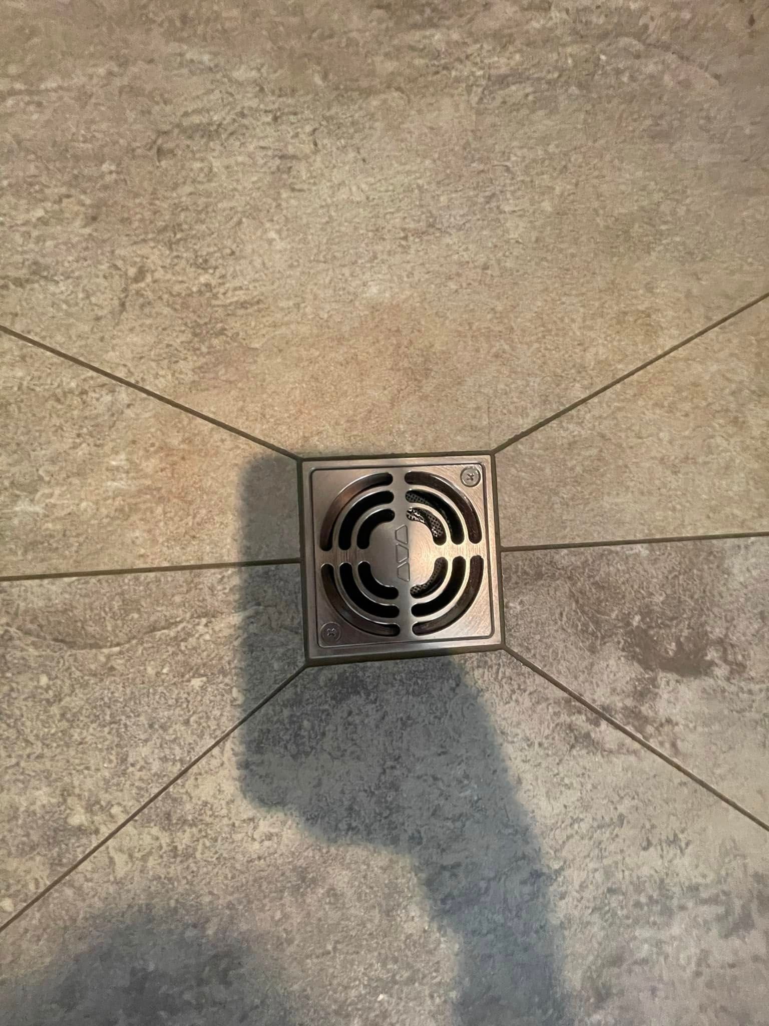 Square shower drain in the center of gray tile floor with black grout lines.