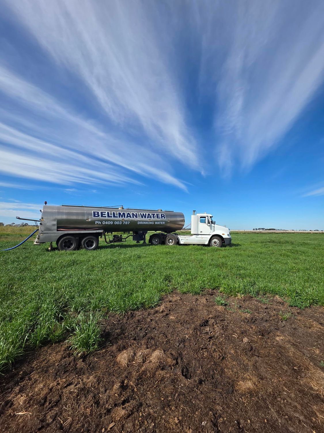 White tanker truck in a green field under a bright blue sky with dramatic clouds