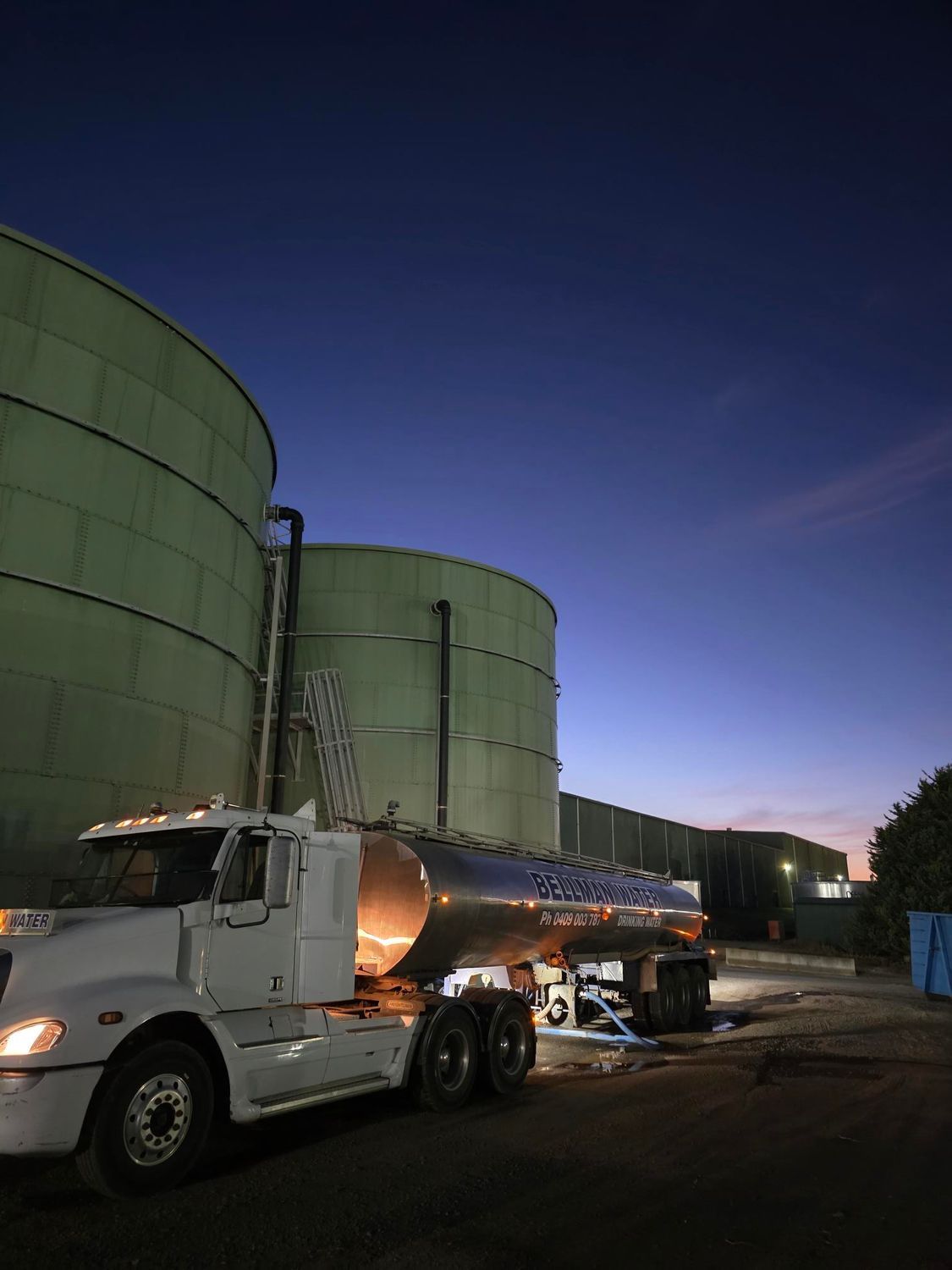 Tank truck parked beside large green storage tanks at dusk