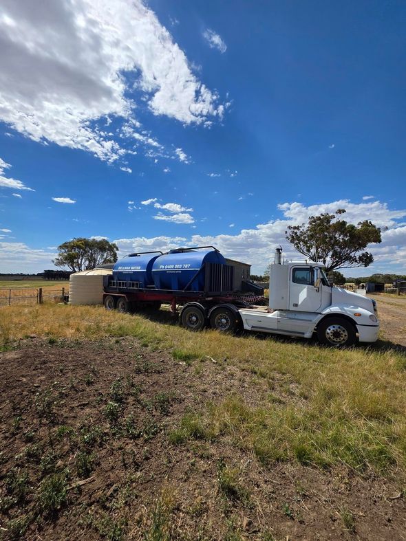 White semi-truck hauling a blue tanker trailer in a grassy field under a bright blue sky