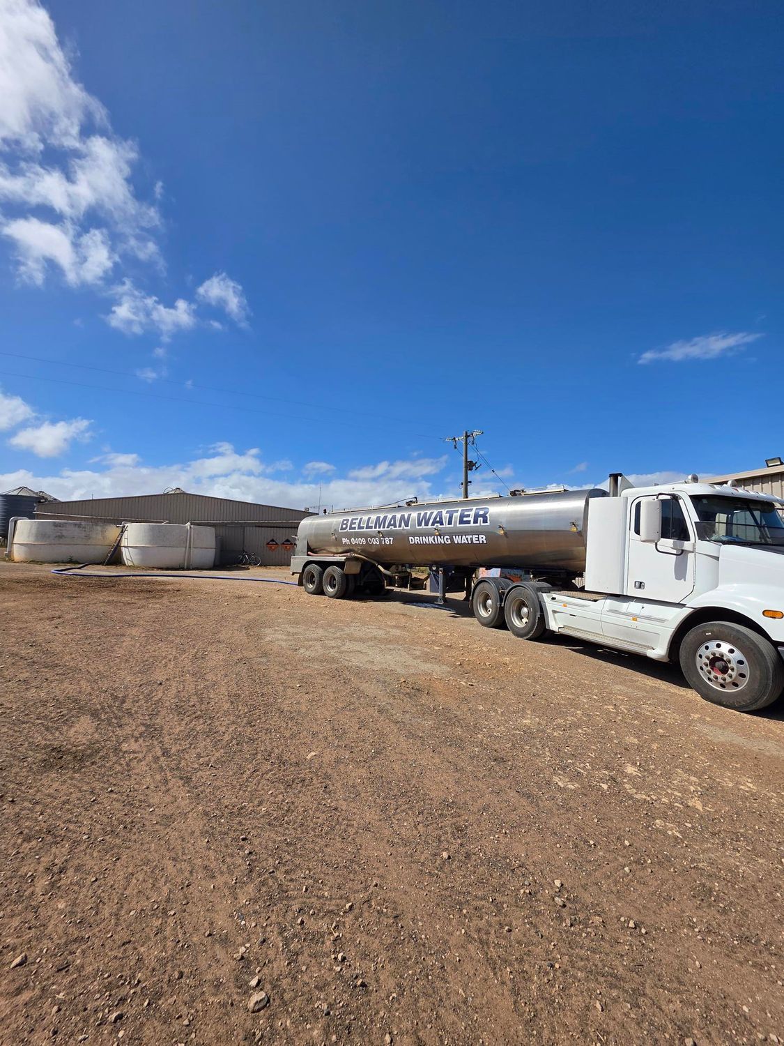 White tanker truck parked on a gravel lot beside large storage tanks under a bright blue sky.