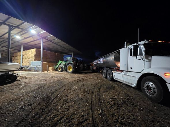 Tractor loading a grain truck beside a barn at night, with a semi-truck in the foreground.