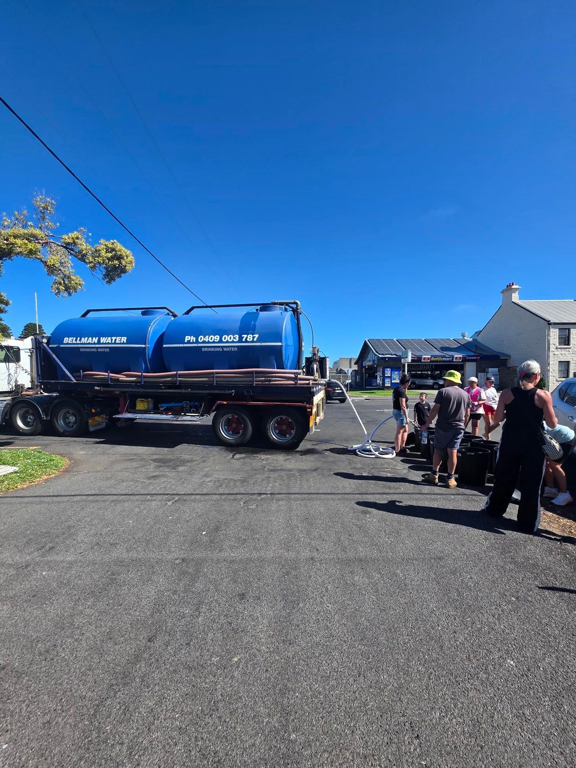 Blue tanker truck on a road with several people standing nearby under a clear blue sky.