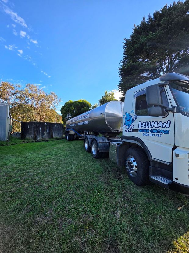 White tanker truck parked on grass under a blue sky beside trees and a small building