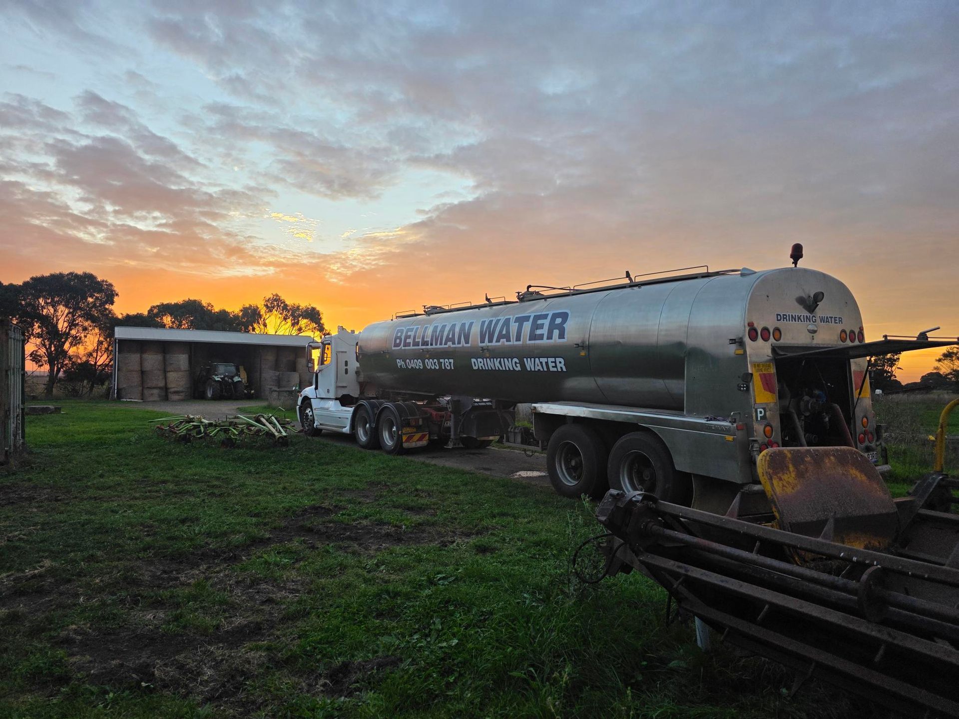 Water tanker truck parked in a grassy yard at sunrise, with a small building and trees in the background