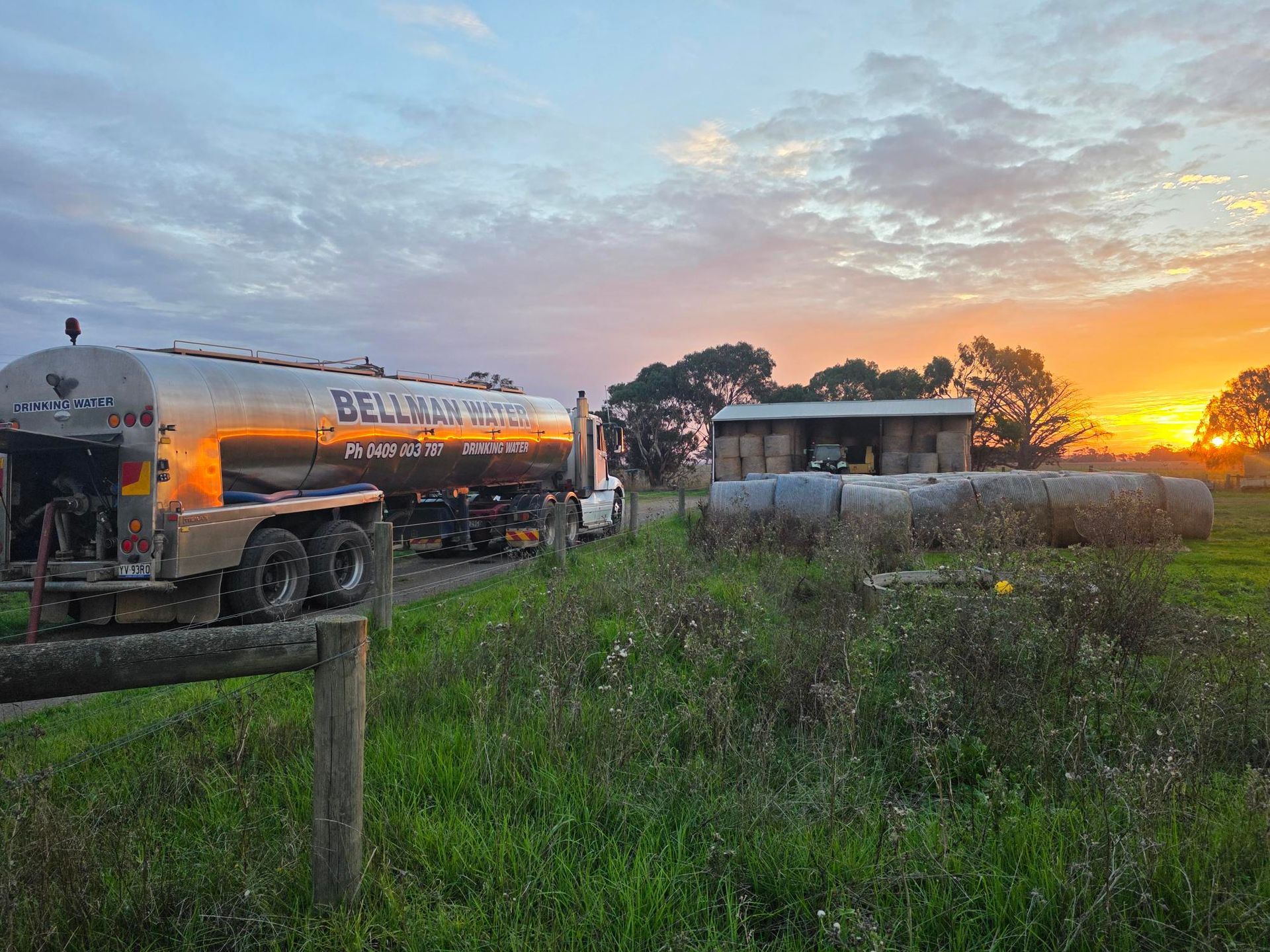 Fuel tanker and small building in a grassy field at sunset, with orange sky on the horizon.