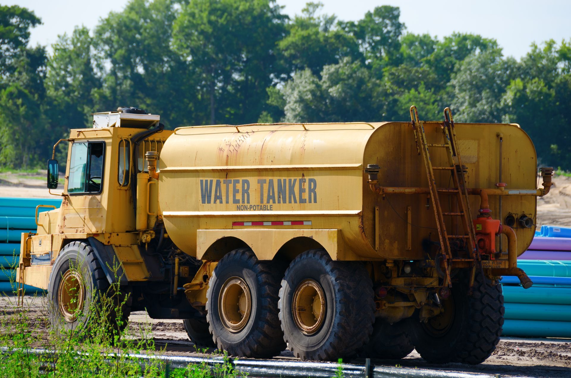 Yellow water tanker truck parked beside blue pipes in a wooded area