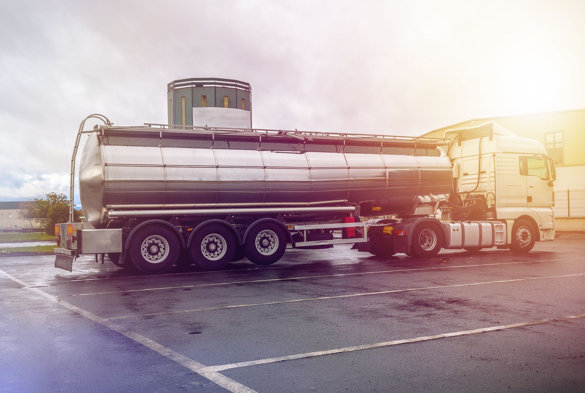 Silver tanker truck parked in a wet lot at sunrise, with sun glare on the right