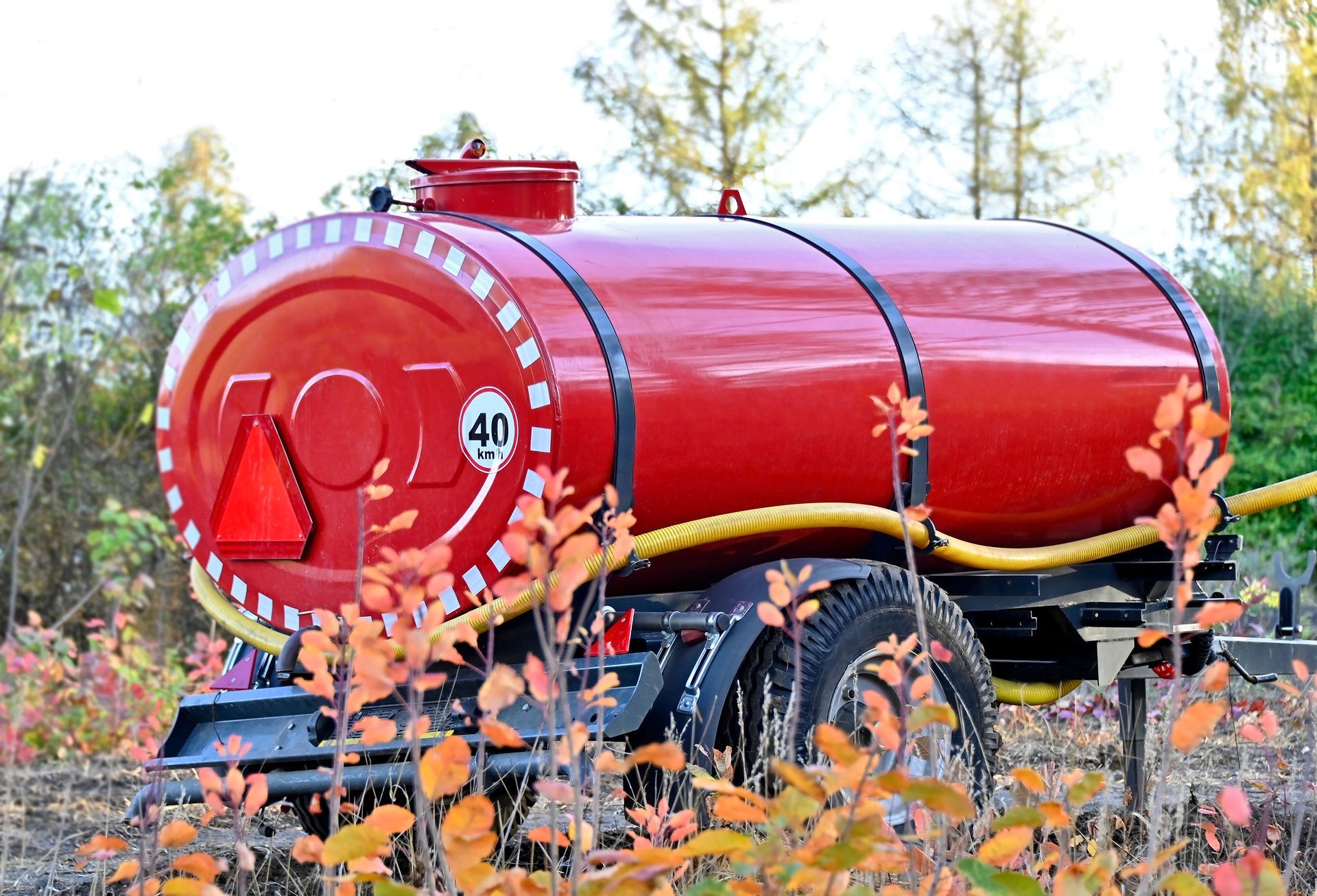 Red cylindrical tanker trailer in a field, bright red with black wheels and autumn foliage in front