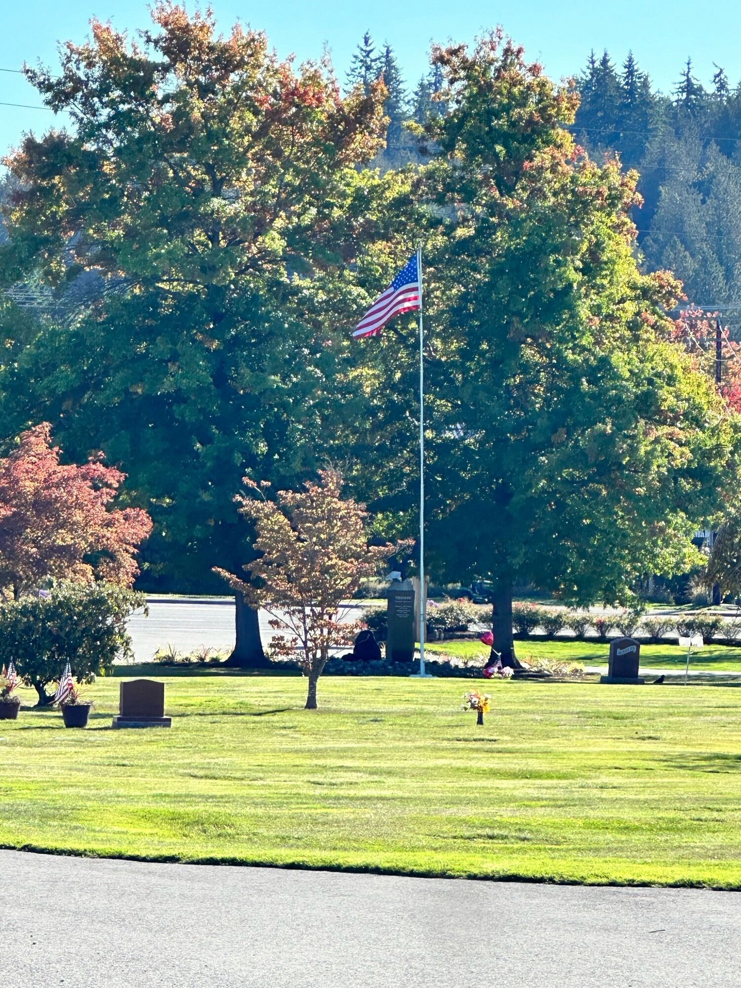 American flag waving in a cemetery with colorful trees and a green lawn under a blue sky.