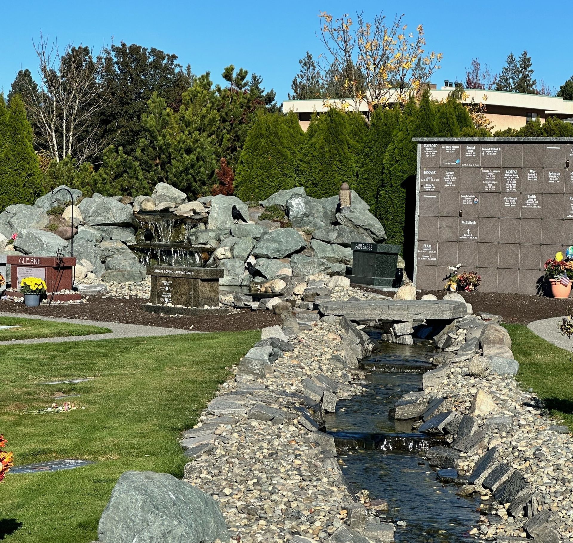 A serene cemetery scene with a water feature, including a small waterfall and stream. Green grass, rocks, and trees.