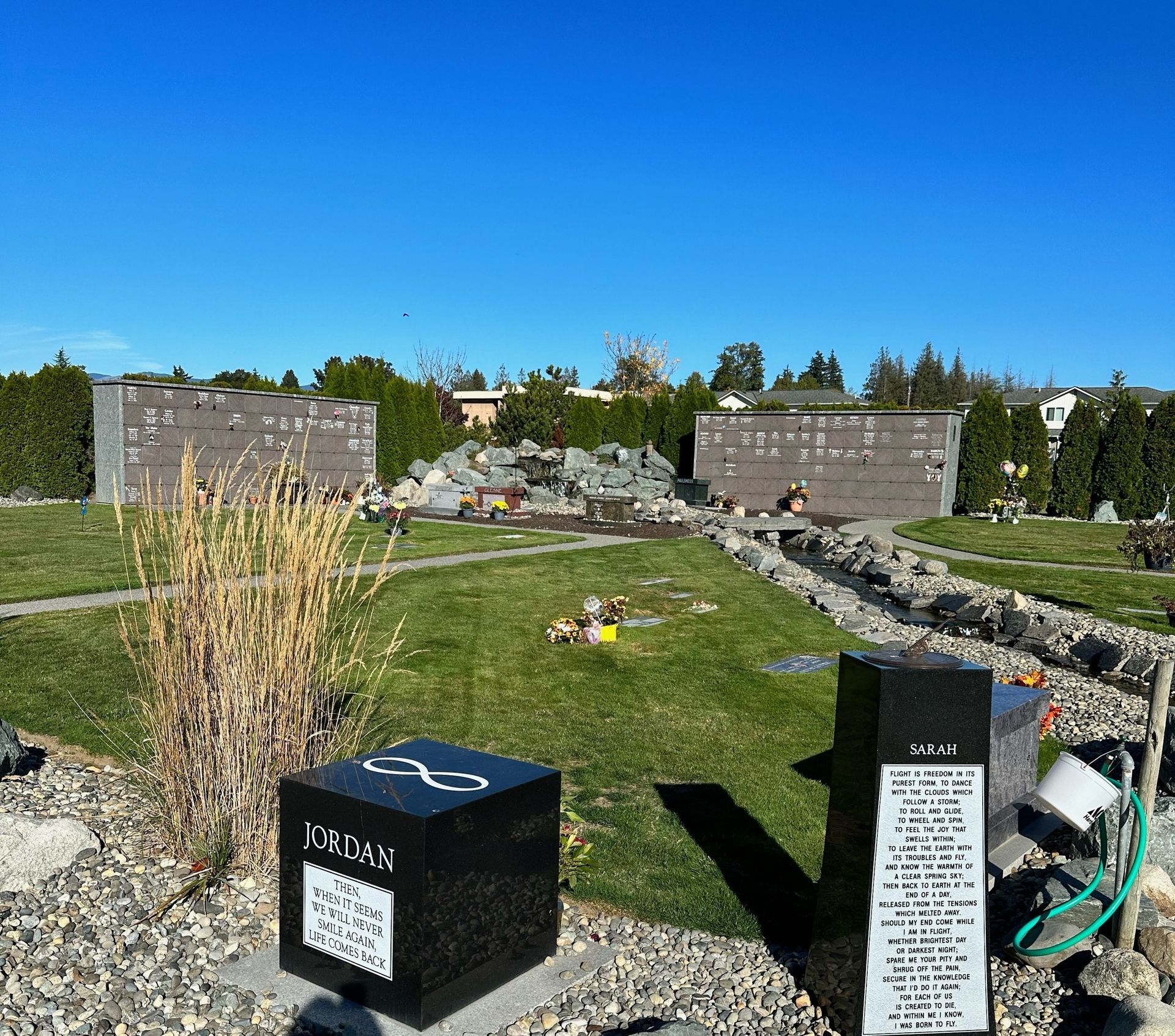 Cemetery scene: black granite tombstones, green grass, two large mausoleums, under a clear blue sky.