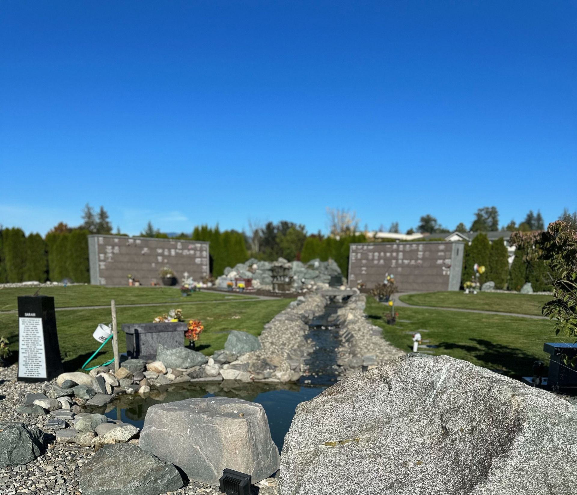 Cemetery with stone memorials, a small water feature, and well-kept lawn under a blue sky.