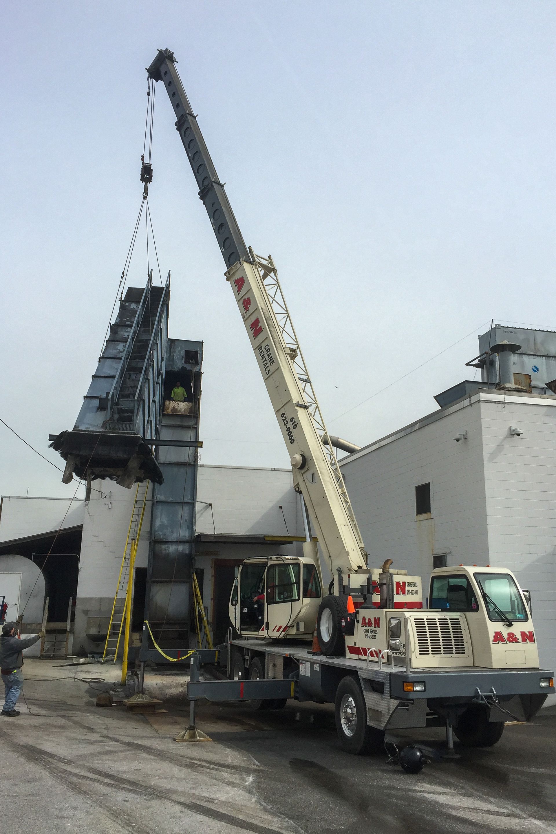 A white A&N Crane Rental crane is parked in front of a building lifting a heavy load. 