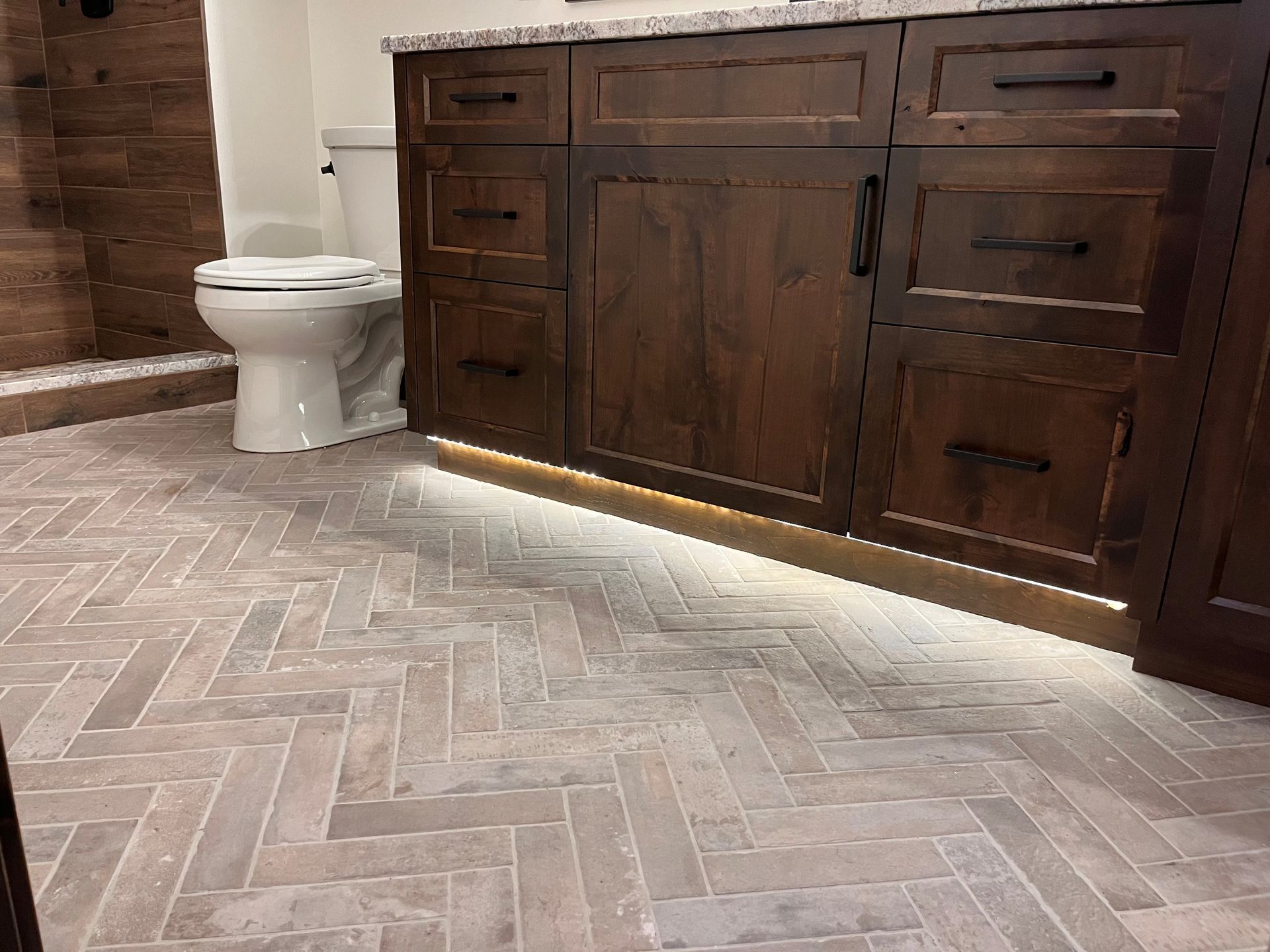Herringbone tile floor in bathroom with dark wooden vanity, toilet, and warm lighting.
