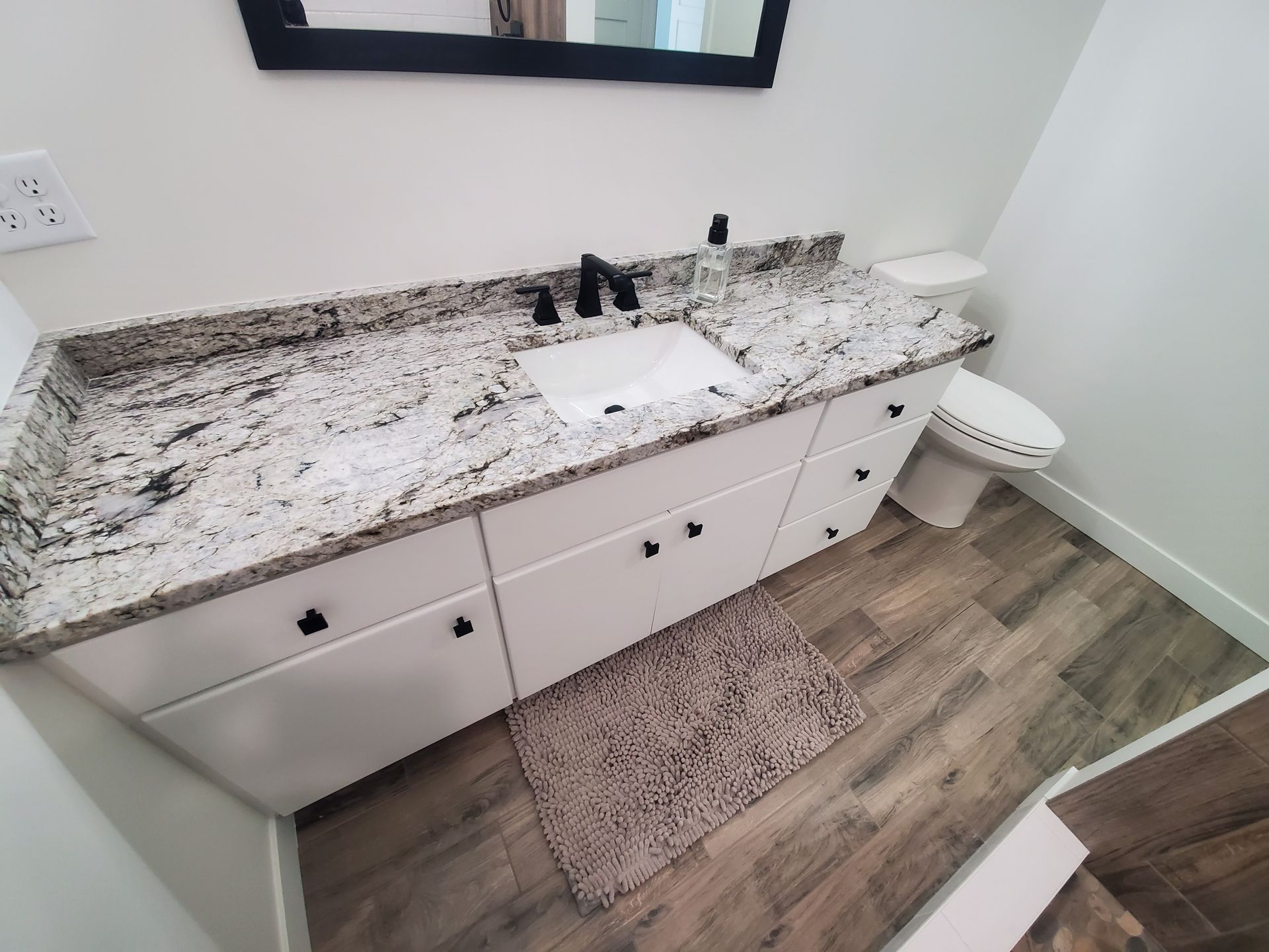 White bathroom vanity with granite countertop, black fixtures, and toilet. Wooden floor and rug.