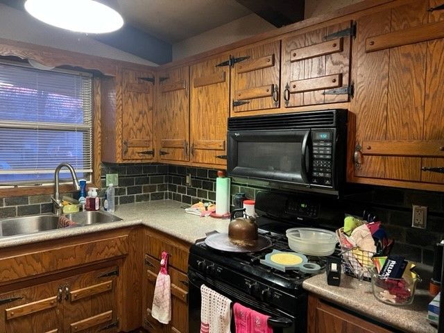 Oak kitchen with black appliances, black backsplash, and cluttered countertops.