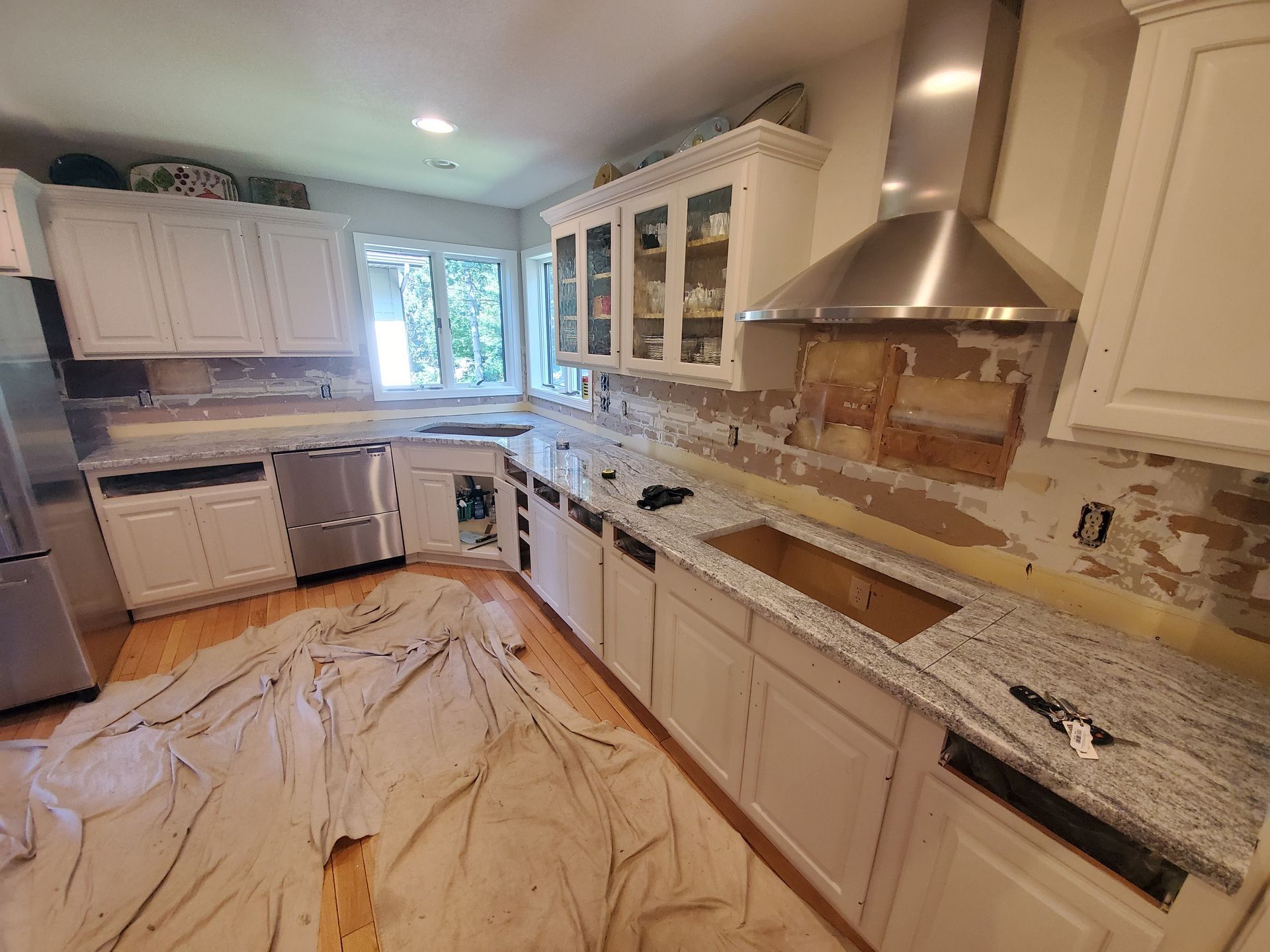 Kitchen renovation in progress: white cabinets, granite countertops, exposed wall, stainless appliances, protective drop cloths.