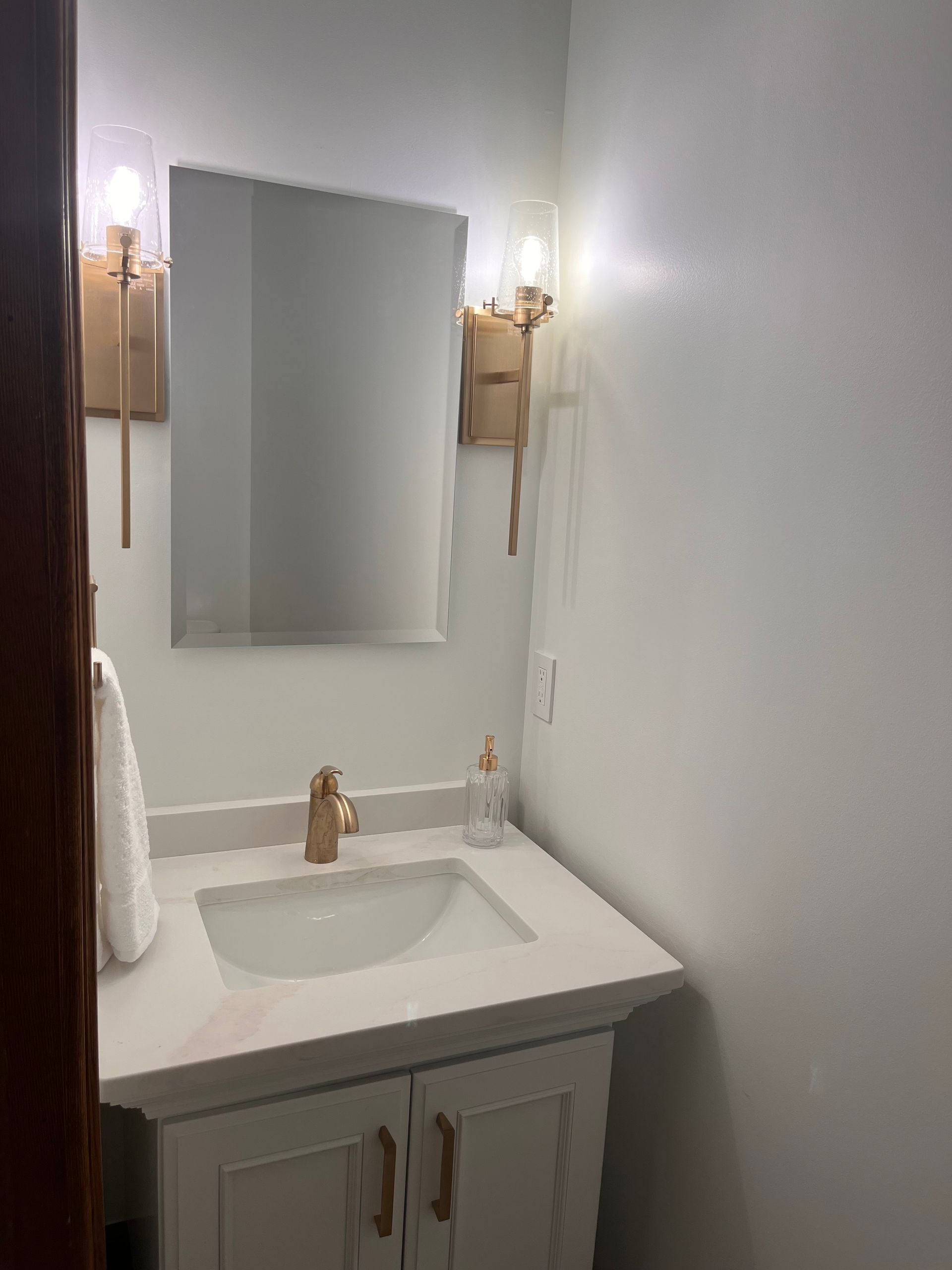 Small bathroom with white walls, vanity, gold faucet and sconces, and a mirror.