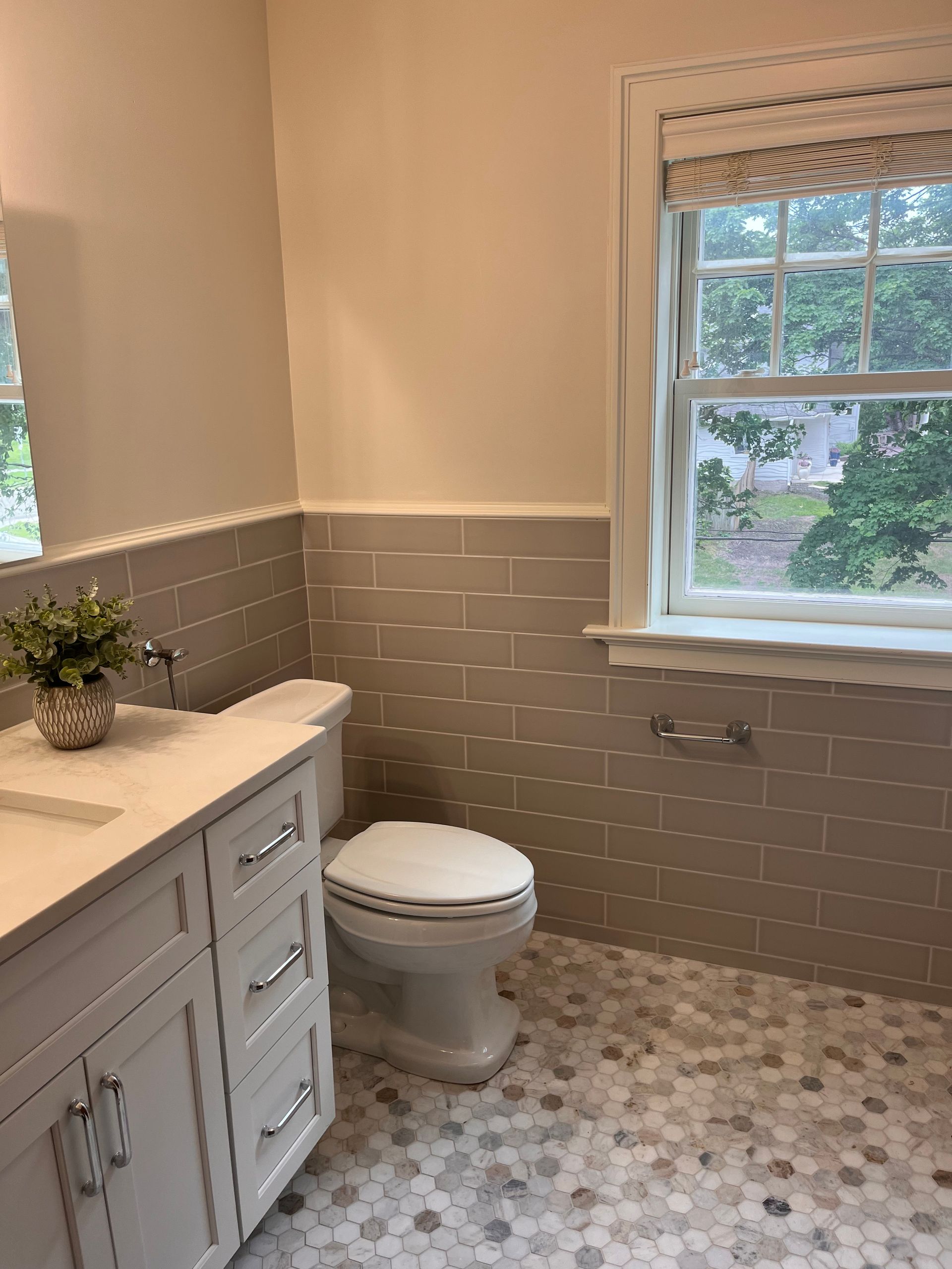 Bathroom with gray tiled walls, white vanity, toilet, and patterned floor, and window with shade.