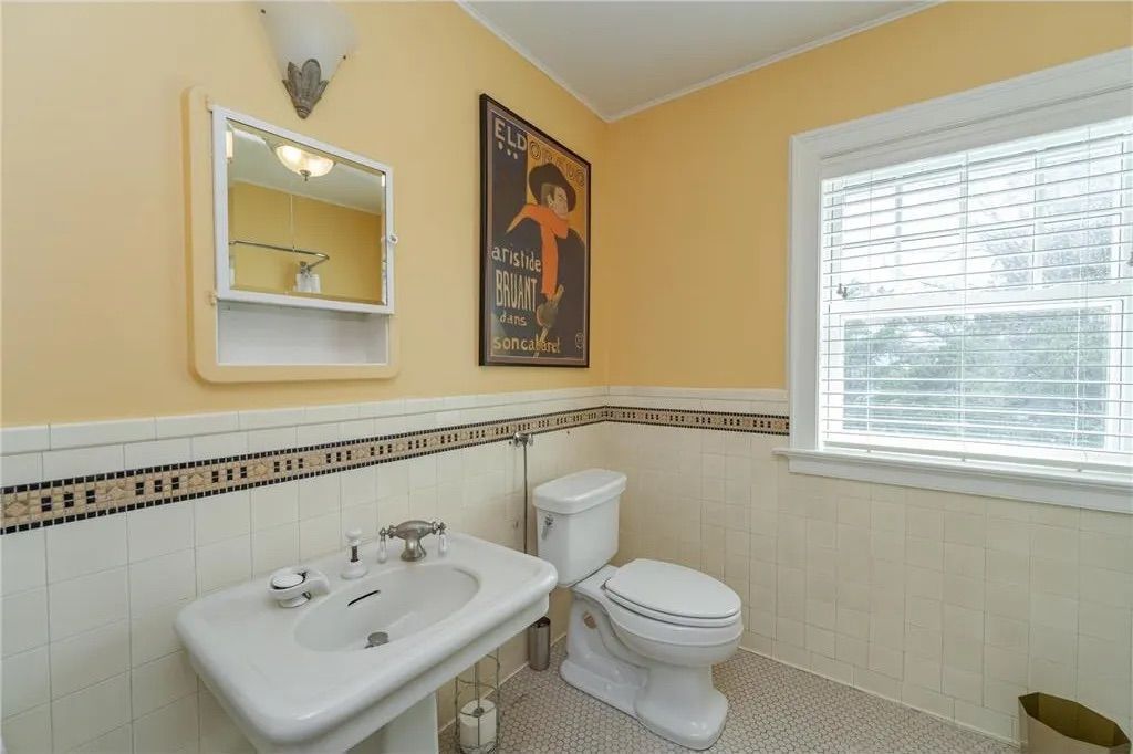 Bathroom with a pedestal sink, toilet, window, and art on the wall. Yellow walls, white tile, and a tiled border.