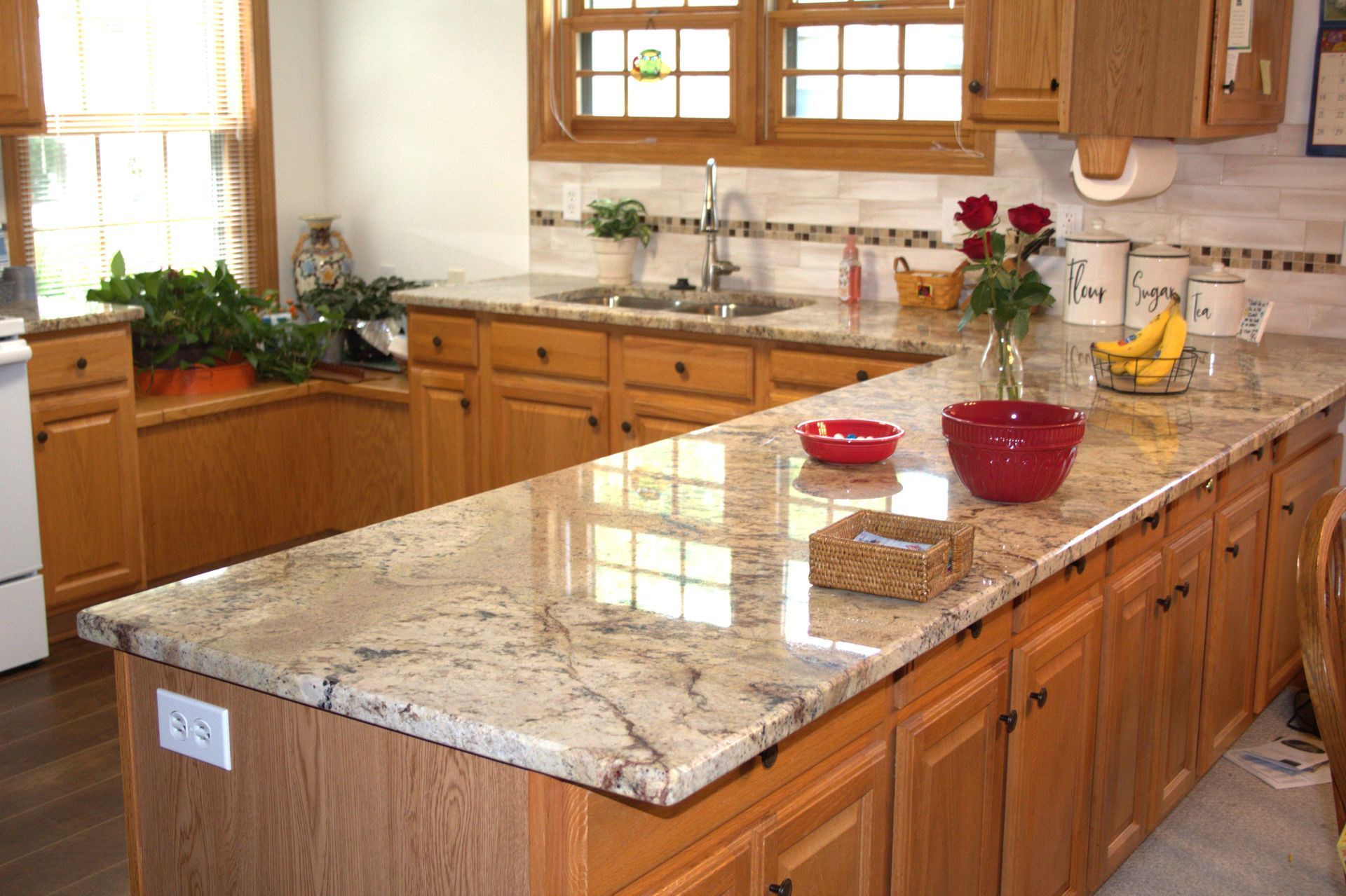 Kitchen with granite countertop, wooden cabinets, sink, window. Red bowls, fruit, and flowers on the counter.