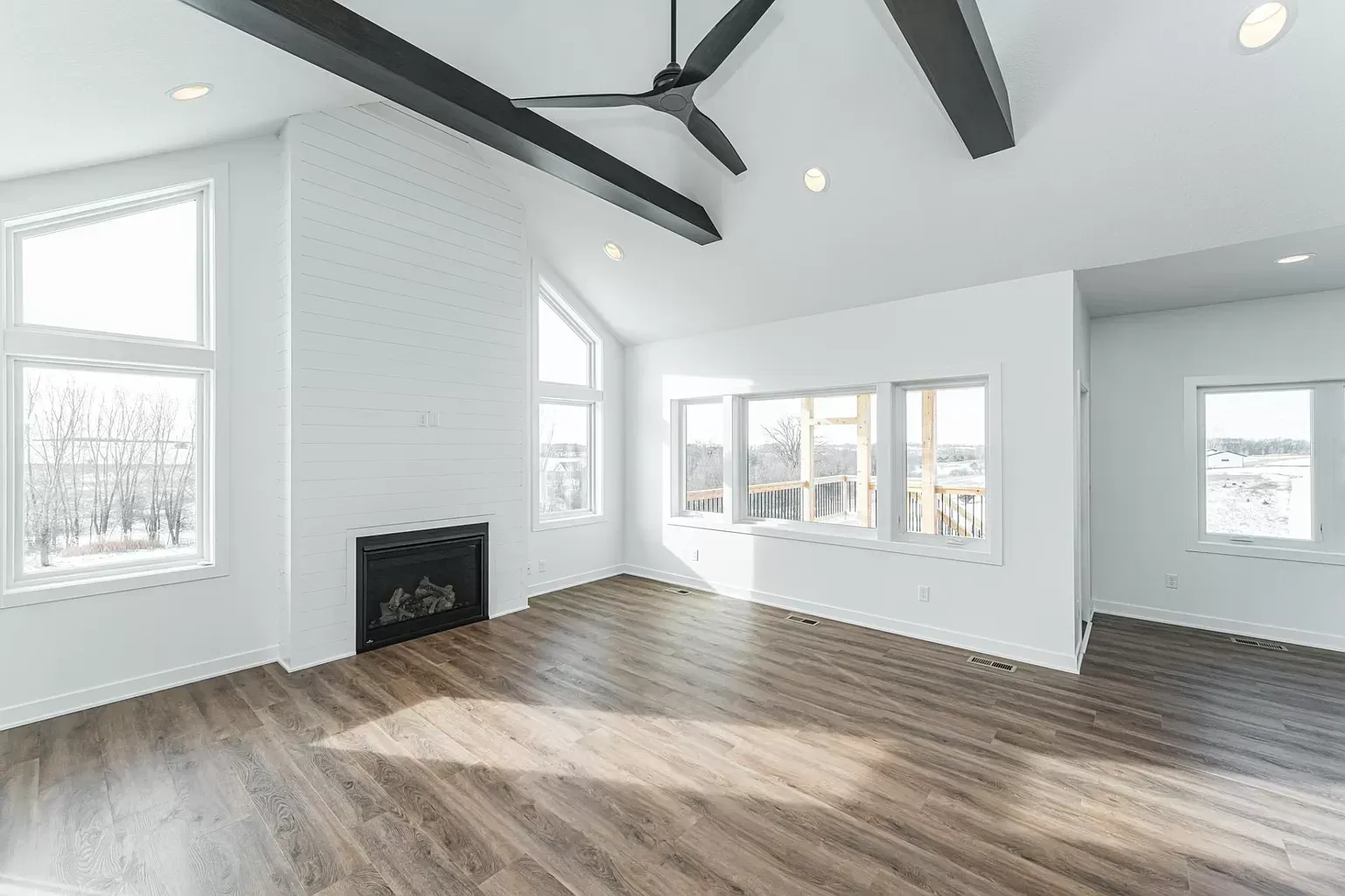 Spacious white living room with fireplace, large windows, and dark wood beams, brown floor.