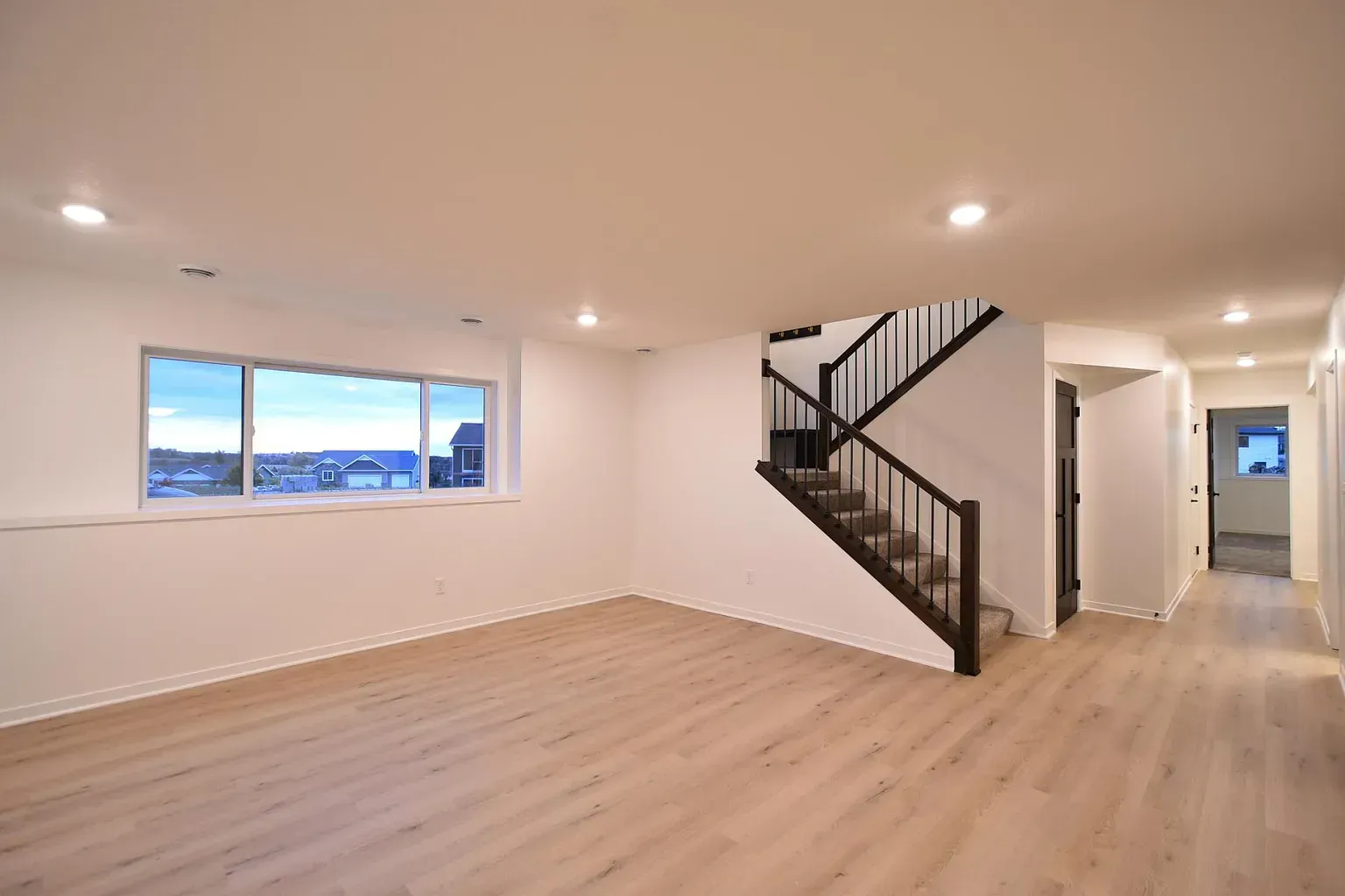 Empty interior with stairs, window, and wood floors. White walls, black railing, and natural light.
