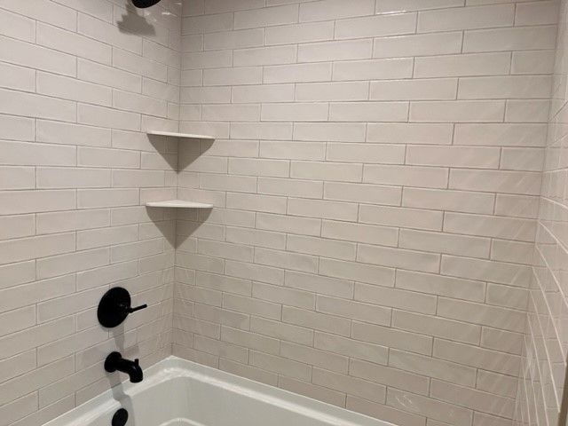 A white-tiled shower with black fixtures, two corner shelves, and a bathtub.