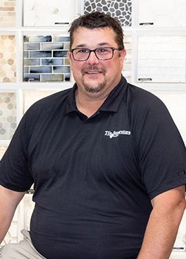 A man wearing glasses and a black shirt is sitting in front of a wall of tiles.