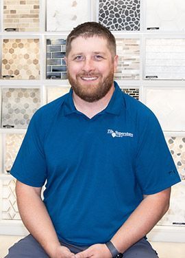 A man with a beard wearing a blue shirt is sitting in front of a wall of tiles.