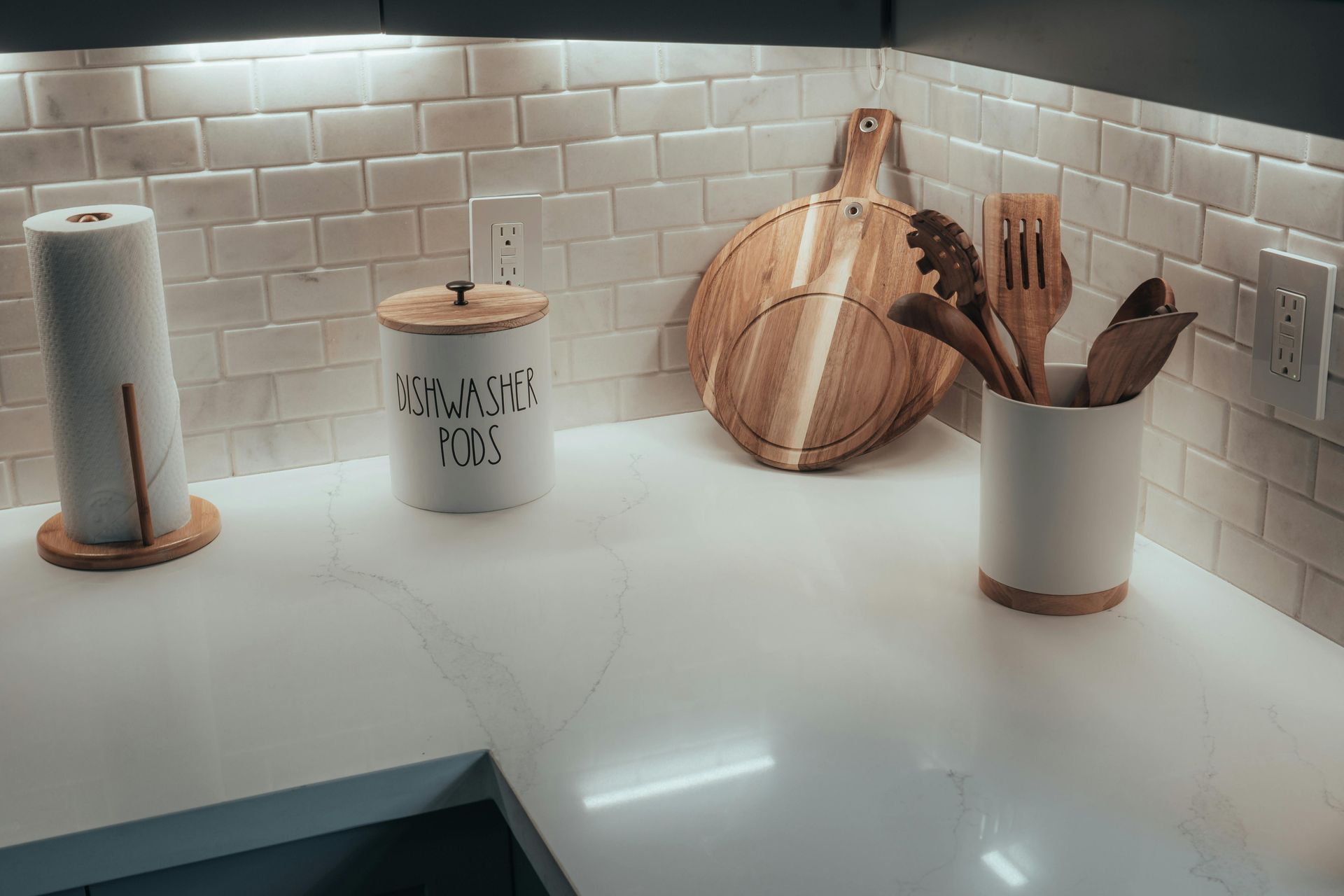 Kitchen countertop with white backsplash, paper towel holder, utensil holder, cutting board, and jars.