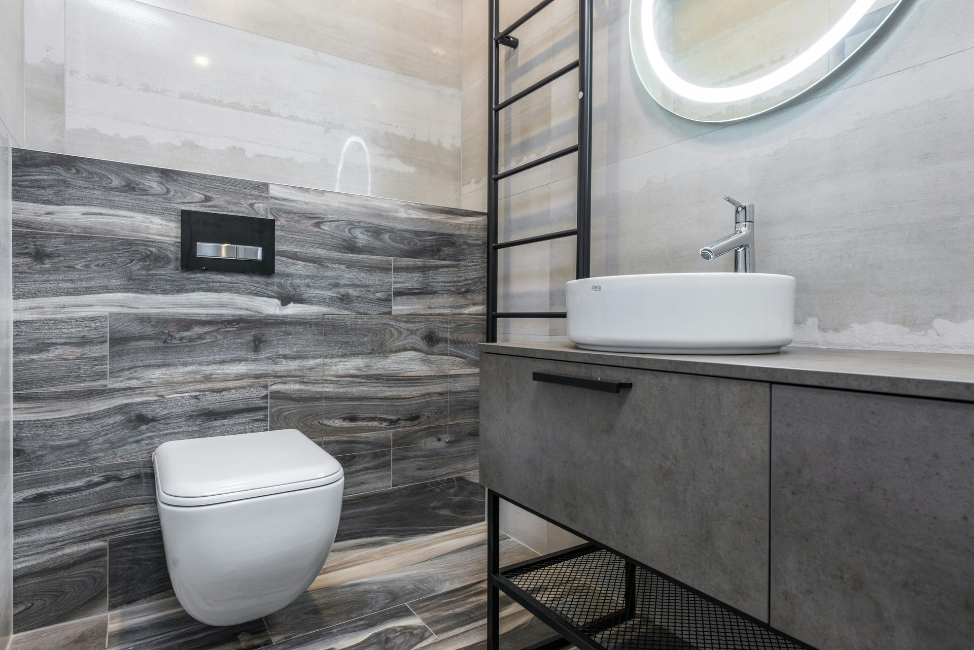 Modern bathroom with gray stone-like walls, floating toilet, gray vanity, white sink, and black ladder-like storage.