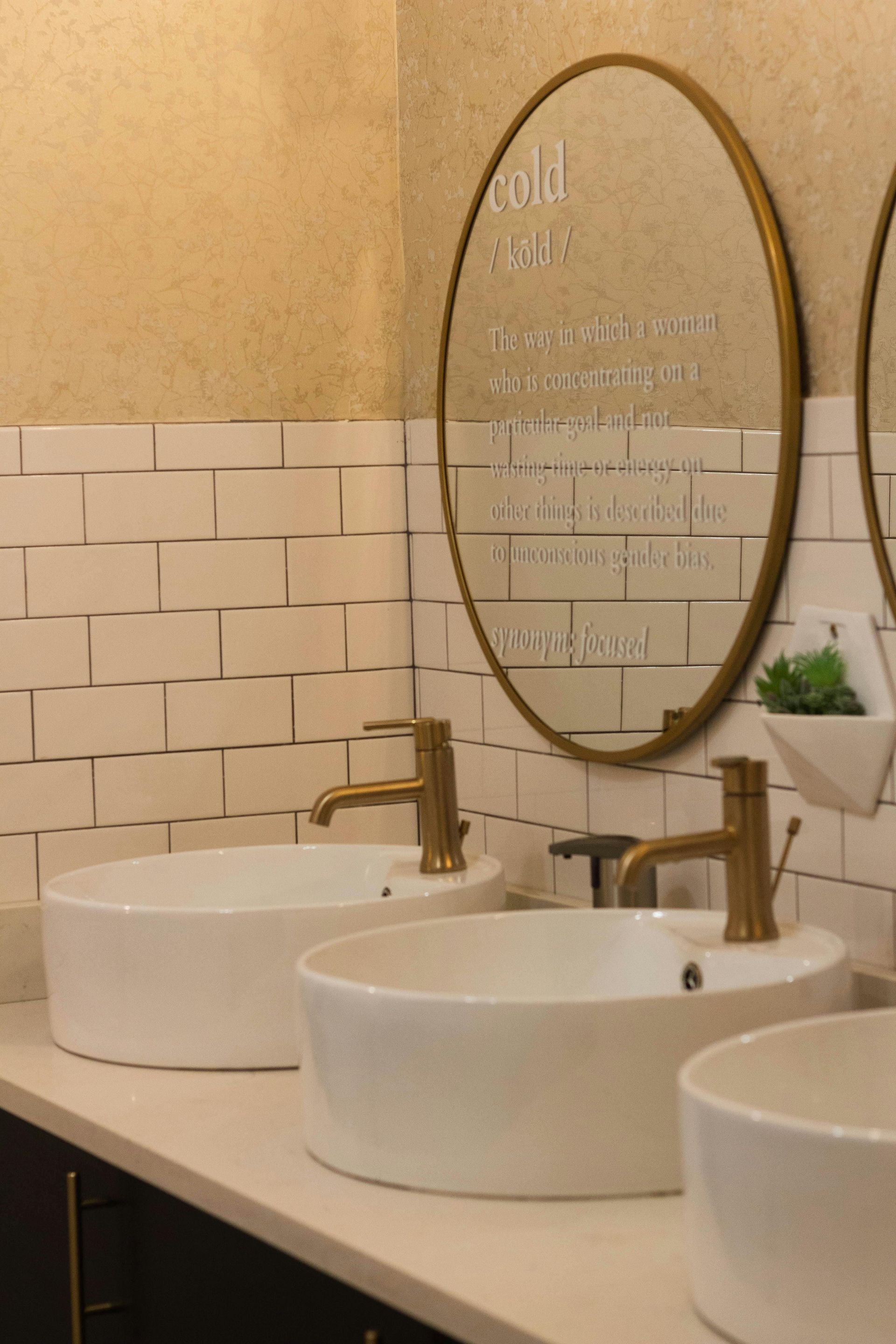 Bathroom with white sinks, gold faucets, and a gold-framed mirror with text; white and gold tile.