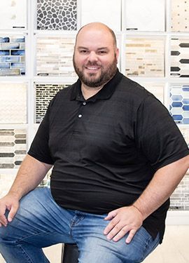 A man with a beard is sitting on a stool in front of a wall of tiles.