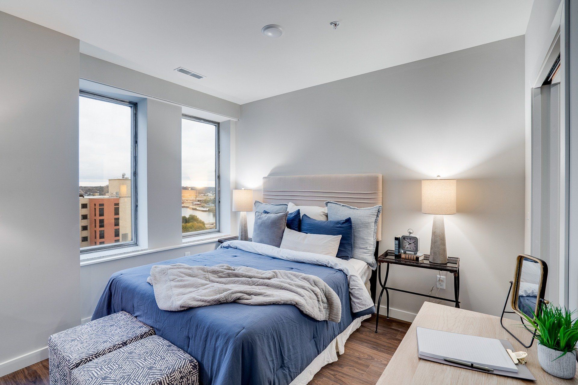 Bedroom in a modern apartment with a blue bedspread, neutral walls, two nightstands, and a large window.