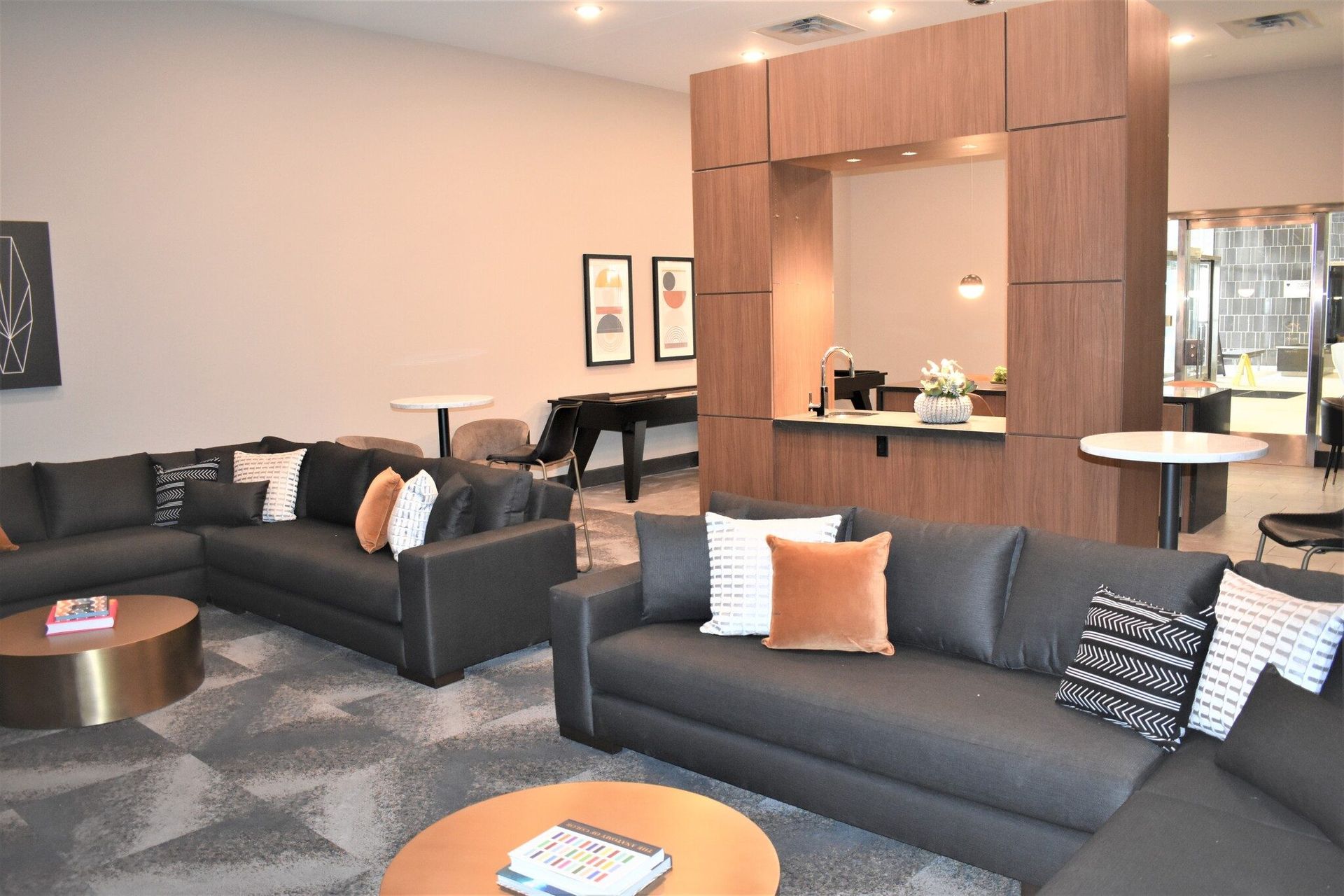 Modern community lounge with dark sectional seating, round coffee tables, and a wood-framed kitchenette area.