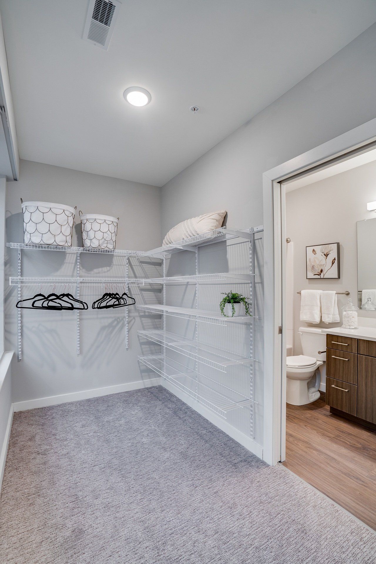 Walk-in closet with white wire shelving, baskets, and hanging rods beside a bathroom.