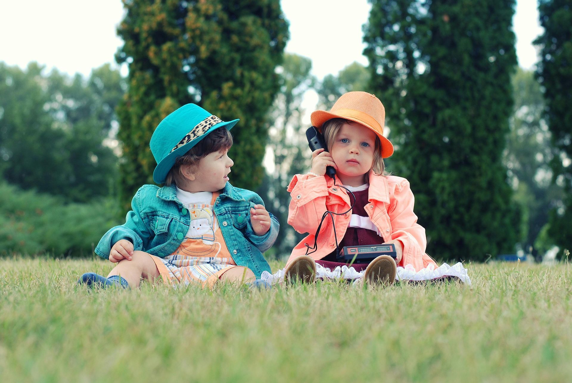 Two children in hats and coats sit on grass; one talks on a phone.