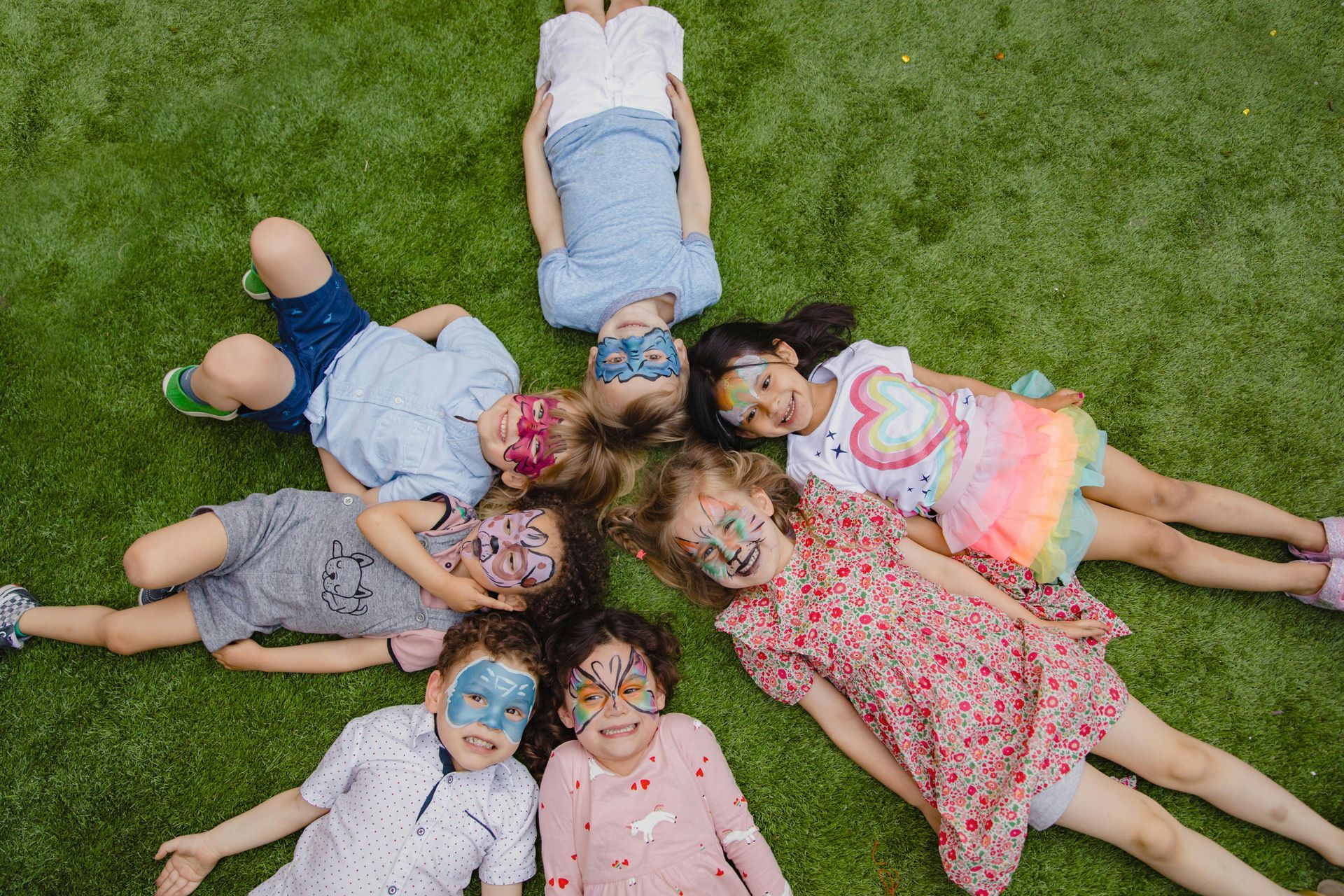 Children with face paint lie in a circle on green grass.