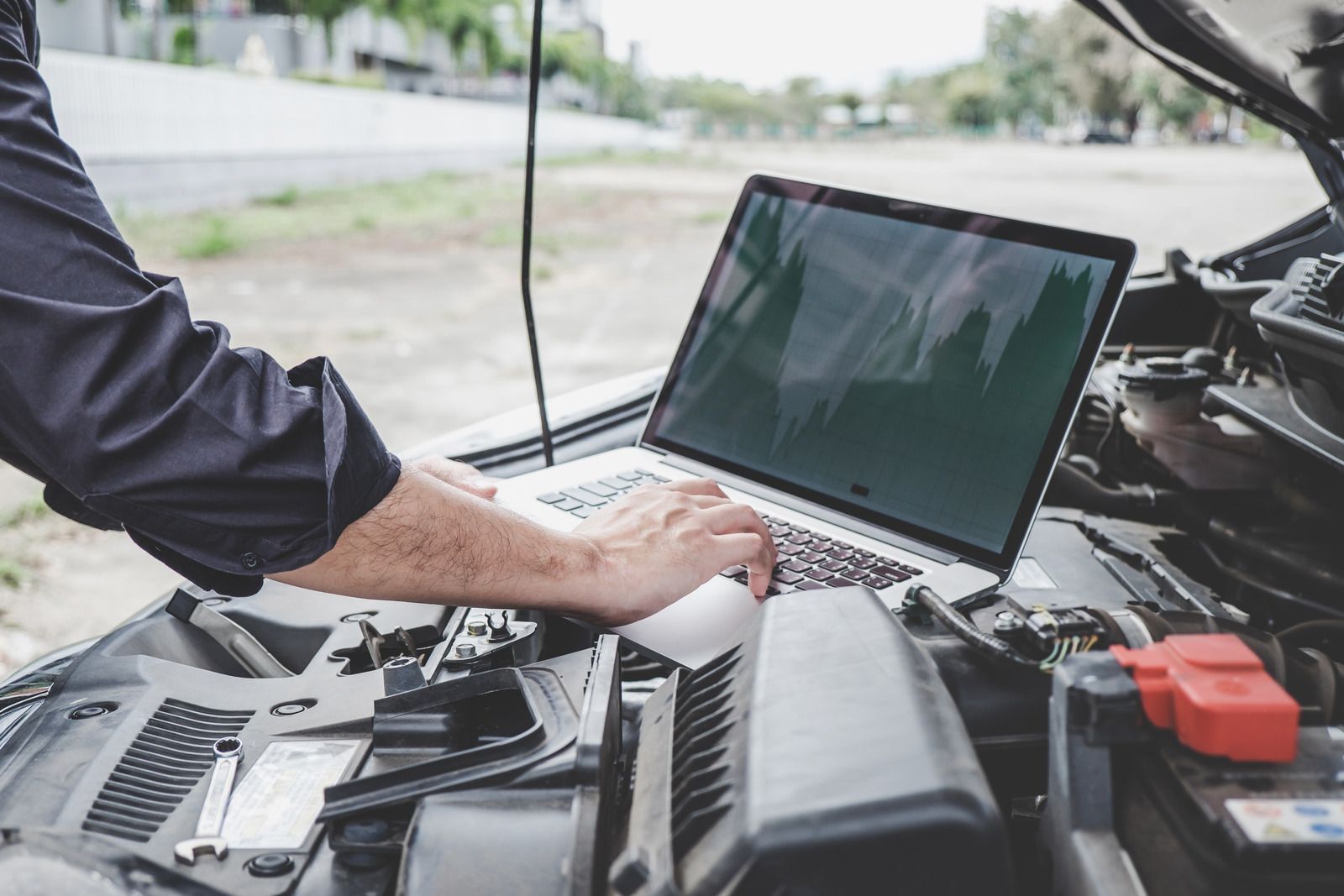 A man is working on a laptop computer under the hood of a car.