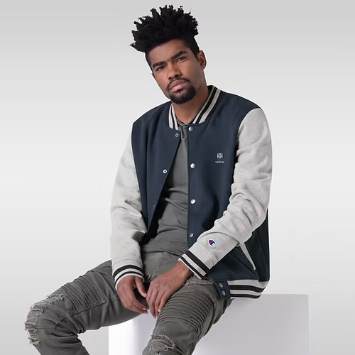A man wearing a baseball jacket is sitting on a white box.